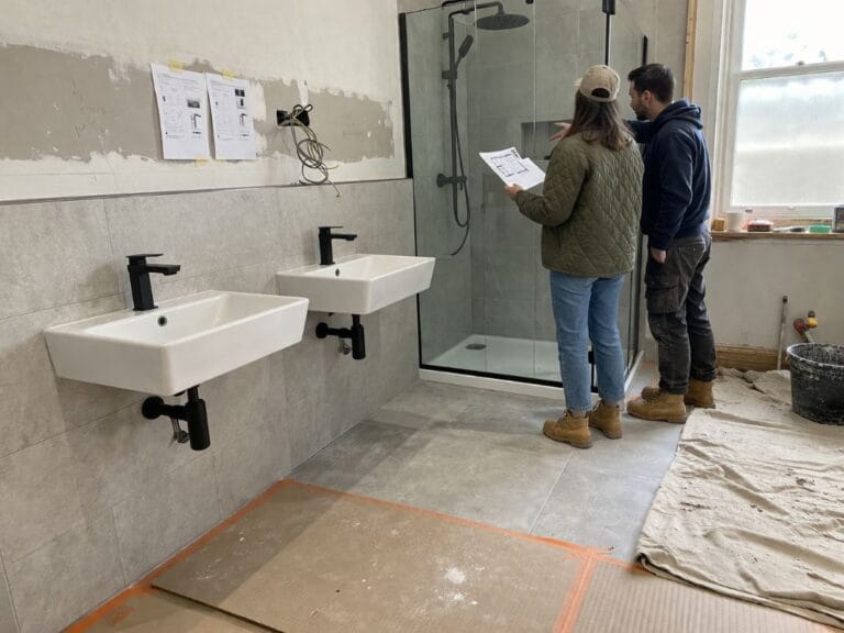 an illustrative image of of modern bathroom fixtures such as wall-hung basins, new taps, and a glass shower enclosure being inspected on a renovation site