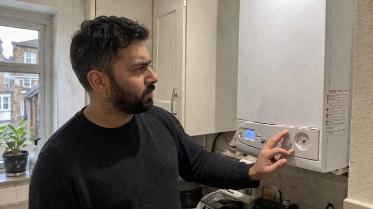 an illustrative image of a man standing in a small London kitchen next to a white wall-mounted boiler, looking at the boiler pressure gauge with a concerned expression
